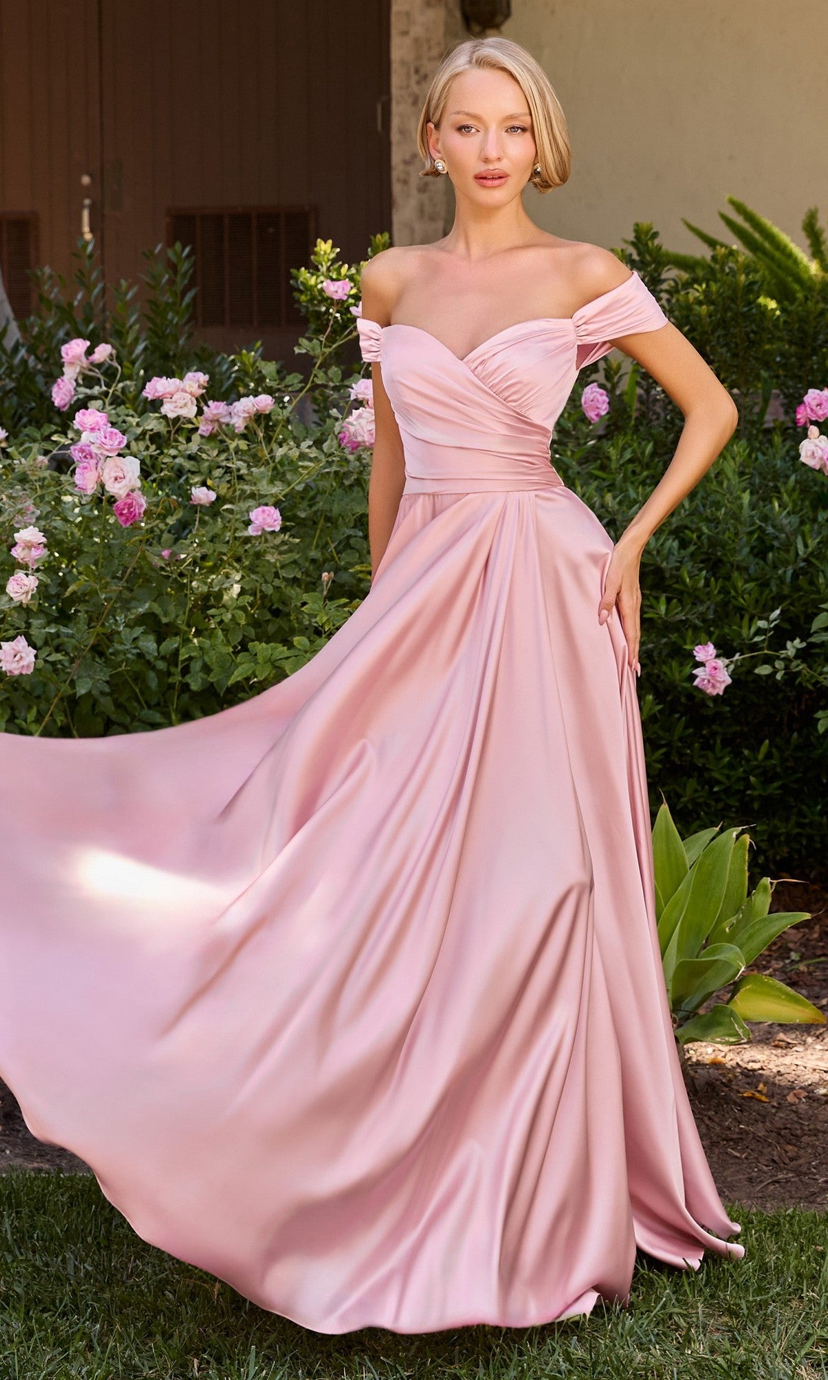 A woman wearing the Classic Off-Shoulder Long A-Line Prom Dress CL7493 stands in a garden surrounded by blooming pink roses and lush greenery.